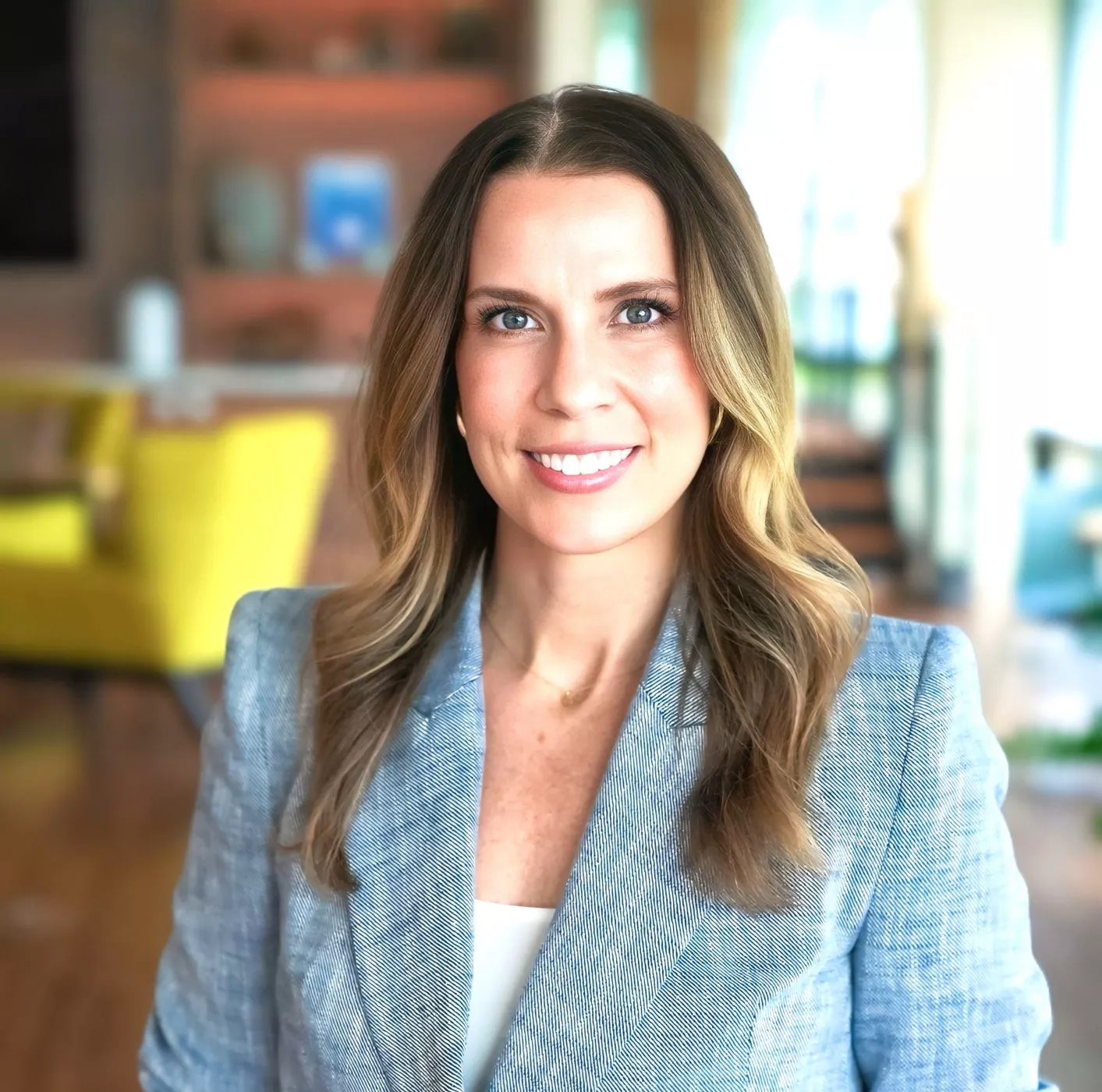 A woman wearing a light blue blazer and white top is positioned in a contemporary office environment. The background features warm tones, modern furniture, and natural lighting through large windows. Her hairstyle is styled in loose waves, complementing the professional attire.